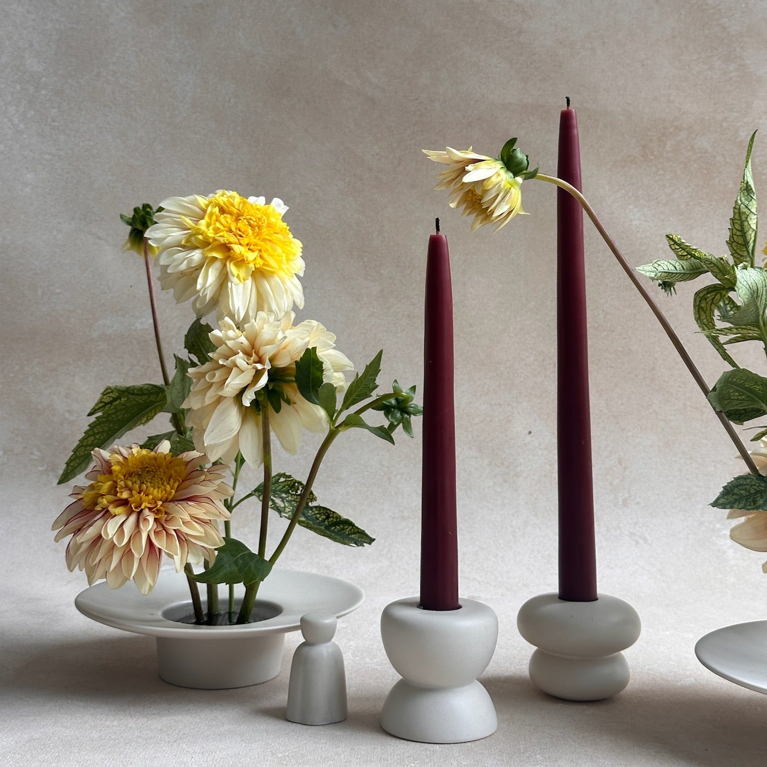 A styled still life featuring two dark burgundy taper candles in white ceramic holders, set beside a white ceramic dish holding cream and yellow dahlias against a textured beige background.
